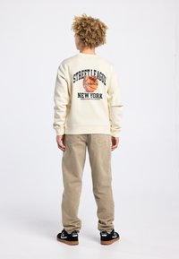 Sweatshirt crème avec un graphique de basket-ball et du texte au dos, associée à un pantalon en velours côtelé beige et des baskets noires avec des accents de logo blancs.