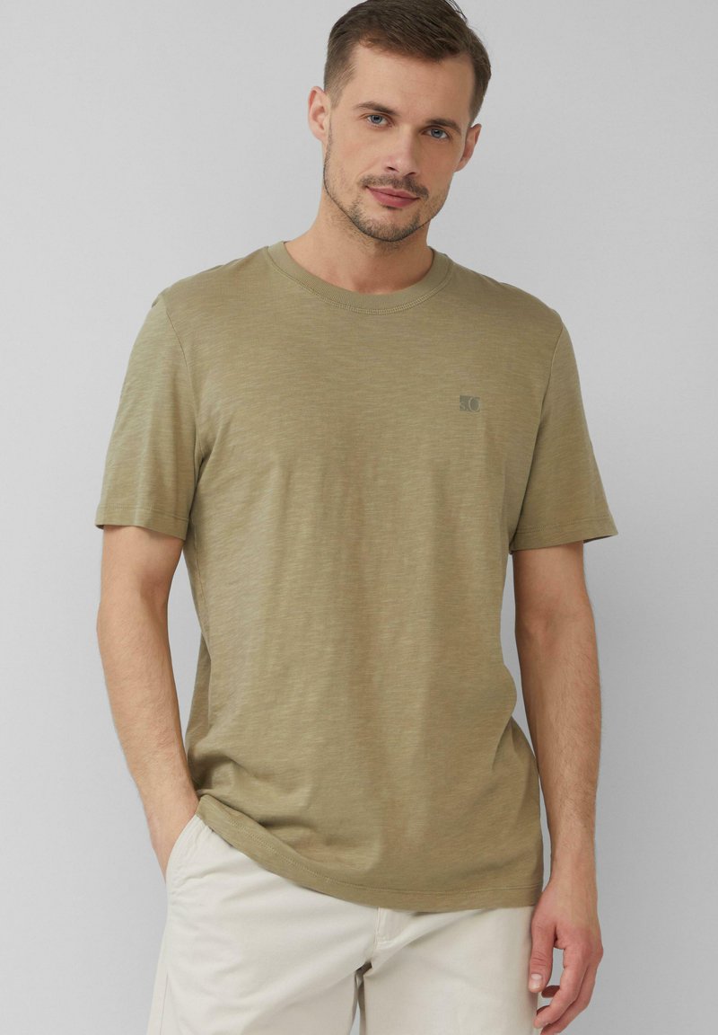 Oliwkowa koszulka z okrągłym dekoltem, wykonana z miękkiego materiału, z krótkimi rękawami i małym logo po lewej stronie klatki piersiowej. Gładka tekstura.
