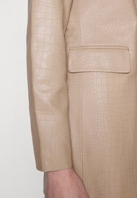 Beige taksinkangladesh nahka takki, jossa on strukturoidun krokotiilinnahkamalli, suora hihansuun malli ja suorakulmainen tasku edessä.