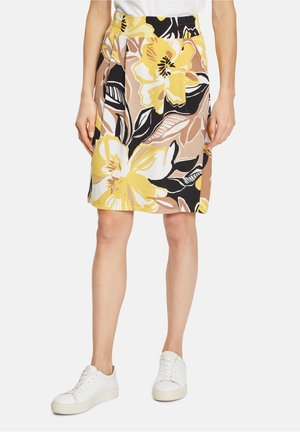 Kvinde iført en knælang nederdel med stort gult, sort, hvidt og beige blomsterprint, kombineret med hvide sneakers.