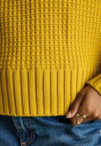 Nahaufnahme einer Hand mit einem goldenen geometrischen Ring, der auf blauem Denim neben dem Saum eines senfgelben Strickpullovers ruht.