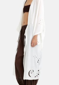 Kobieta w białym kimono z rozcięciami w kwiatowe wzory, nosząca brązowy top bandeau i luźne spodnie, stoi na tle białym.