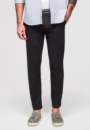 Mann trägt schwarze Slim-Fit-Hose, graue Tassel-Loafers, ein weißes Hemd und ein hellblaues offenes Hemd mit Knöpfen, Hände in den Taschen.
