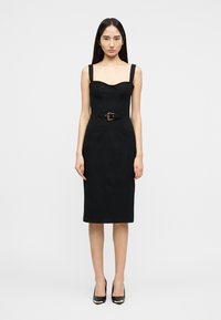 Γυναίκα που φοράει εφαρμοστό μαύρο φόρεμα μέχρι το γόνατο με φαρδιές τιράντες, ζώνη με διακοσμητική αγκράφα και μαύρα ψηλοτάκουνα παπούτσια, όρθια.