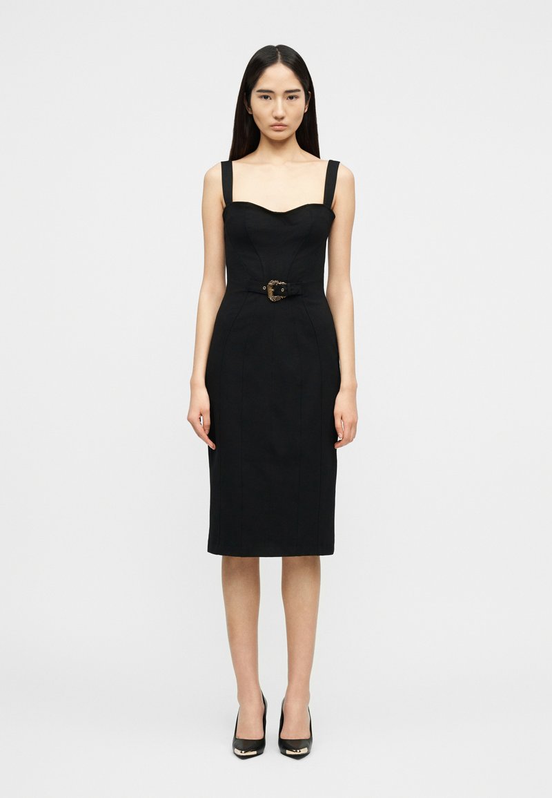 Γυναίκα που φοράει εφαρμοστό μαύρο φόρεμα μέχρι το γόνατο με φαρδιές τιράντες, ζώνη με διακοσμητική αγκράφα και μαύρα ψηλοτάκουνα παπούτσια, όρθια.