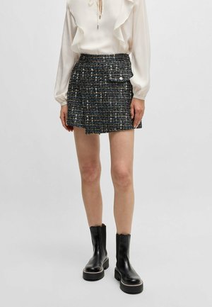 Frau, die eine weiße Bluse mit Rüschen, einen schwarzen Tweed-Minirock mit Fronttasche und schwarze Plateau-Stiefeletten vor einem einfarbigen Hintergrund trägt.