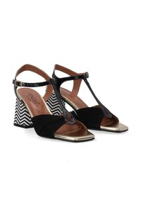 Schwarze Riemchensandalen mit patentiertem Lederobermaterial, goldenen Akzenten und einem blockförmigen Absatz mit Chevron-Muster. Offene Zehen- und Knöchelriemen-Design.