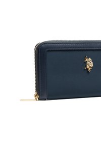 Granatowy skórzany portfel z złotym akcentem w postaci logo, zamykany na zamek błyskawiczny, o fakturowanej powierzchni. Cechuje się prostymi liniami i prostokątnym kształtem.