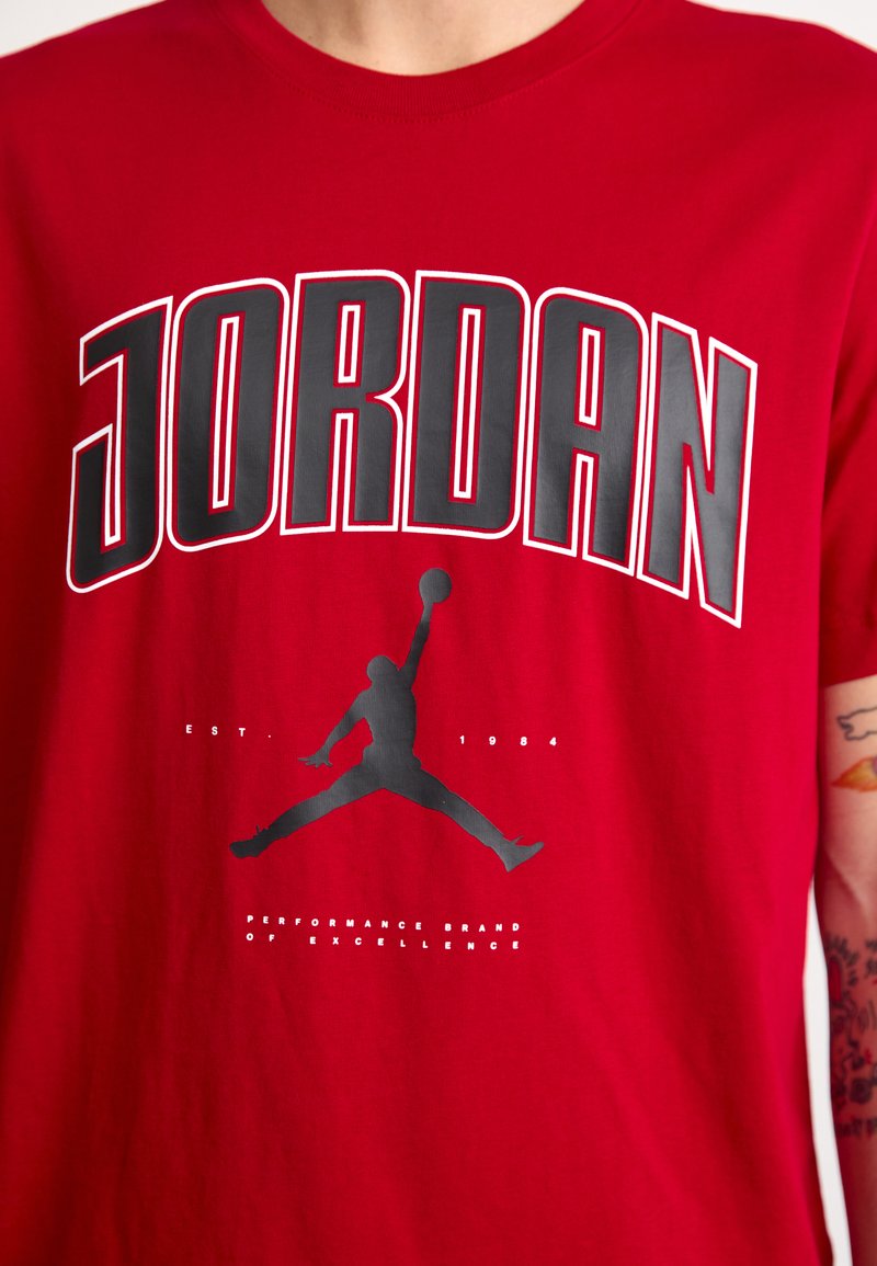 Rotes Baumwoll-T-Shirt mit fettgedrucktem schwarzen Text "JORDAN", einer schwarzen Basketball-Silhouette und "EST. 1984" in glatter Textur.