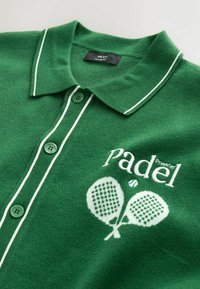 Polo vert avec passepoil blanc, boutons et texte brodé « Padel Premier » au-dessus de deux raquettes de padel croisées et une balle.