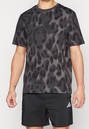 Mężczyzna w czarnej i ciemnoszarej, wzorzystej koszulce Adidas oraz czarnych szortach Adidas stoi z rozluźnionymi ramionami wzdłuż ciała.
