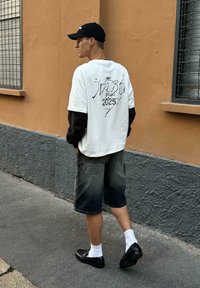 Bílý tričko s černými dlouhými rukávy, s stylizovaným textem na zádech, spárované s tmavými džínovými šortkami a černými botami.