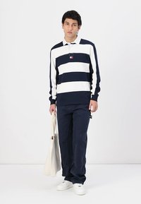 Marineblauwe en witte gestreepte lange mouwen polo met kraag, gecombineerd met marineblauwe broek en witte sneakers. Houdt een beige draagtas vast.