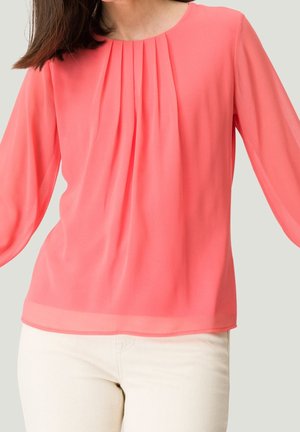 Kvinde iført en koral lyserød bluse med lange ærmer og forreste læg samt hvide bukser, stående mod en ensfarvet lys baggrund.