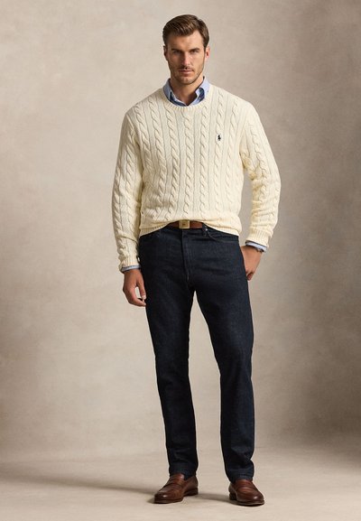 Uomo che indossa un maglione lavorato a treccia color crema sopra una camicia blu, jeans scuri, cintura marrone e scarpe marroni, in piedi davanti a uno sfondo neutro.