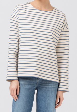 Personne portant une chemise crème à manches longues avec de fines rayures horizontales bleues et un jean en denim bleu, debout devant un fond uni.