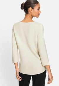 Cremefarbenes Strickoberteil mit Dreiviertelärmeln, das einen runden Halsausschnitt und einen leicht geschwungenen Saum hat, getragen über schwarzen Hosen.