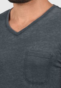 Dunkelgraues V-Ausschnitt-T-Shirt aus weichem Stoff, mit einer kleinen Tasche auf der linken Brust und einem schlichten, gesäumten Rand.