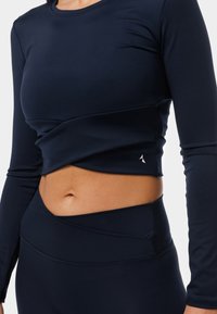 Tume sinise värvi pikkade varrukatega sportlik särk, millel on keha lähedal istuv disain, koos keeratud esiküljega ja väikese logoga allääres. Siledast, venivast kangast.