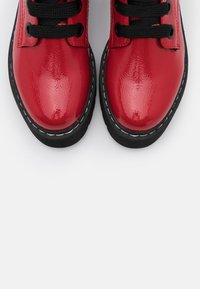 Scarpe in vernice rossa con lacci neri, dotate di punta tonda e cuciture bianche lungo la suola. Superficie liscia e lucida.