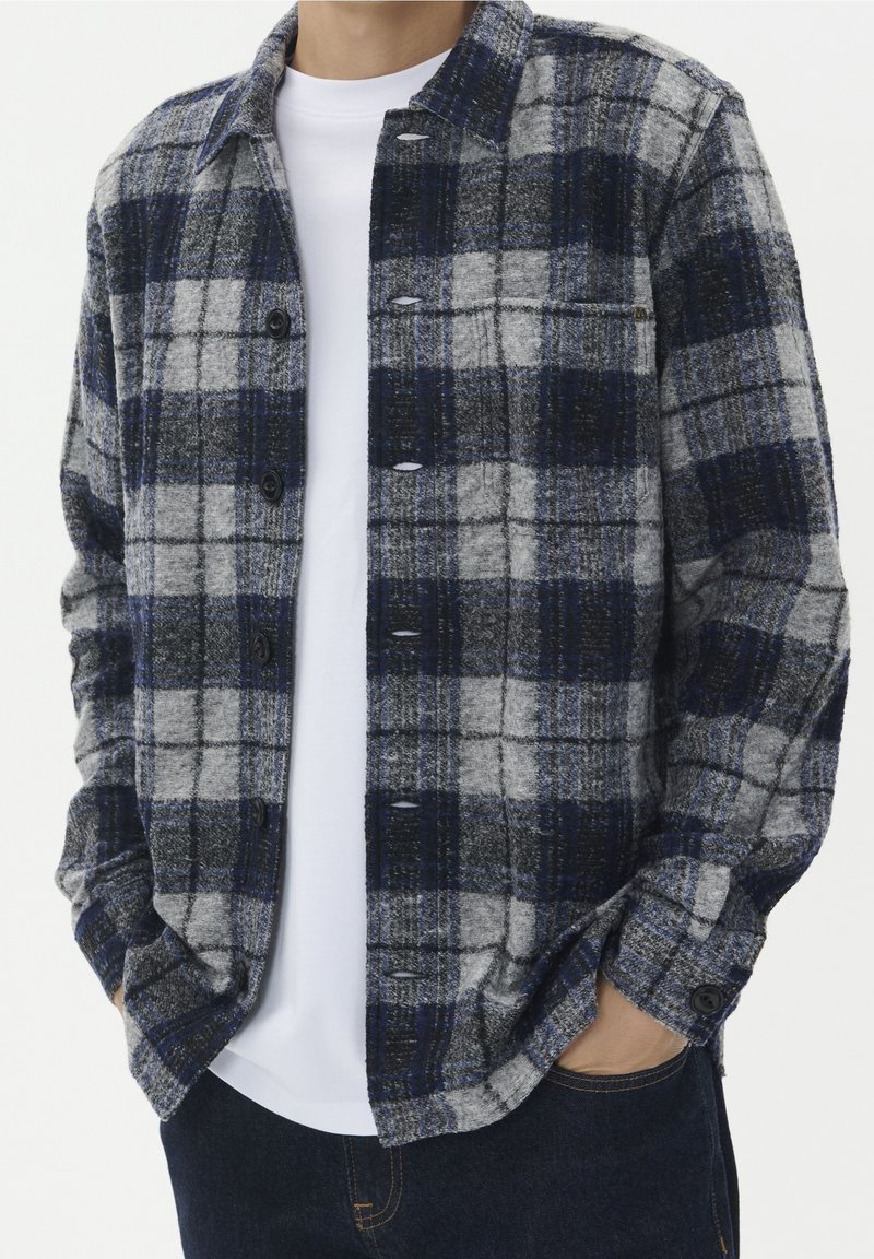 Personne portant une chemise à carreaux bleue, grise et noire boutonnée par-dessus un t-shirt blanc, mains dans les poches d'un jean foncé.