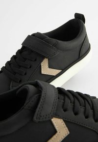 Černé tenisky vyrobené z hladké kůže, s páskem na suchý zip, černými tkaničkami, bílou gumovou podrážkou a béžovým zigzagovým akcentem po straně.