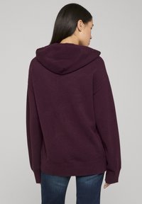 Sweat à capuche bordeaux avec une coupe décontractée, un ourlet côtelé et une texture douce. Présente des épaules tombantes et une capuche spacieuse.