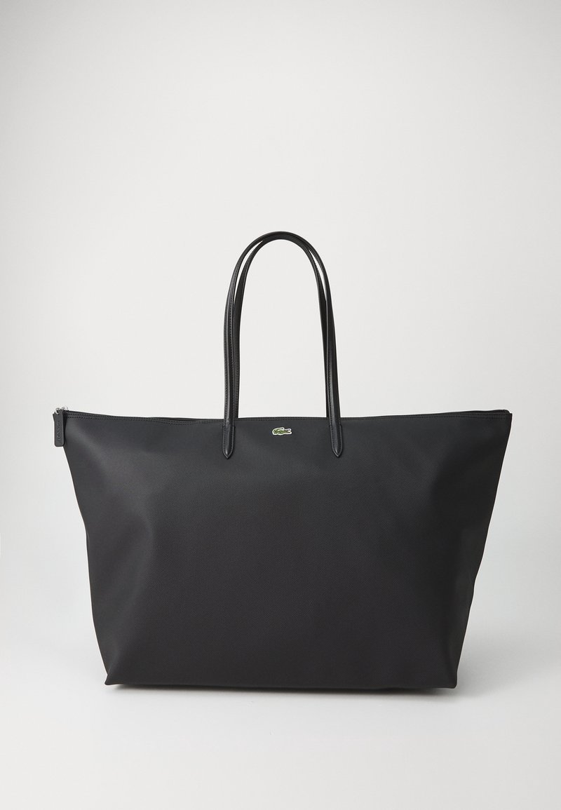Grand sac cabas noir avec deux anses supérieures et un petit logo crocodile vert sur le devant, sur un fond clair uni.
