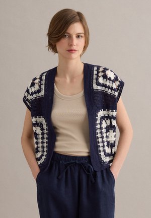 Giovane donna che indossa un top smanicato beige e un gilet all'uncinetto blu navy con motivi quadrati, abbinato a pantaloni navy con coulisse, mani nelle tasche.