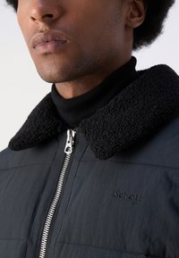 Sort pufferjakke med en struktureret sort fleecekrave. Har en sølvfarvet lynlås og broderet logo på brystet. Glat stofstruktur.