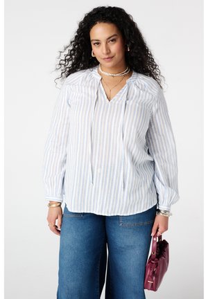 Vrouw met krullend haar, draagt een gestreept wit-blauw blouse en jeans, met een kleine bordeauxrode handtas in haar rechterhand.