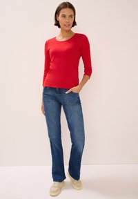 Roter Langarmoberteil mit rundem Ausschnitt, kombiniert mit blauen Schlagjeans und hellen Sneakers. Einfaches Design, glatte Textur.