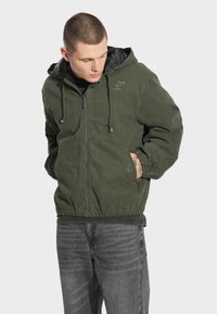 Zielona kurtka z kapturem z zamkiem z przodu, elastycznymi mankietami i zewnętrznym materiałem. Posiada czarną pikowaną podszewkę oraz logo na piersi.