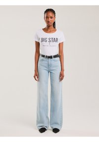 Naine punutud juustega, kellel on seljas valge "BIG STAR" t-särk, mis on sisse toppitud helesinistesse kõrge vöökohaga laiade säärega teksadesse, millel on must vöö ja mustad kingad.