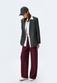 Blazer gris de doble botonadura, camisa blanca, pantalones de pierna ancha burdeos y gorra gris. Los zapatos son blancos, con una textura suave.