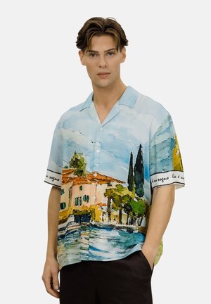 Ung mann iført en kortermet skjorte med et naturskjønt akvarellmotiv av hus, trær og vann, poserer med den ene hånden i lomma.