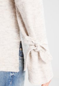 Lichtbeige trui met een zachte textuur, voorzien van een strikdetail aan de mouw en een geribbelde zoom, gecombineerd met blauwe denim.