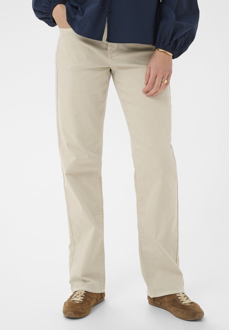 Persona che indossa pantaloni beige a gamba dritta, sneakers marroni con stringhe, maglia blu scuro a maniche lunghe, con una mano in tasca e gioielli dorati sulla mano destra.