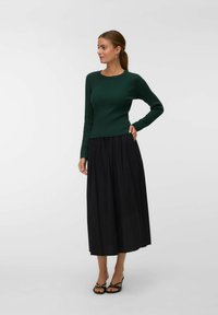 Femme aux cheveux bruns en queue de cheval, portant un pull côtelé vert foncé, une jupe midi noire, des sandales à talons noires et des boucles d'oreilles en argent.