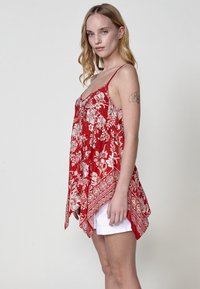 Tunique sans manches rouge avec des motifs floraux blancs, décolleté en V et ourlet asymétrique. Associée à un short blanc, doté de fines bretelles.