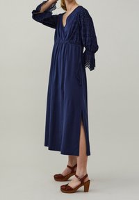 Tummansininen maxi-mekko, jossa on syvä V-kaula-aukko ja pitsiset puffihihat. Mekossa on joustava vyötärö ja sivuhalkiot. Yhdistetty ruskeisiin korkokenkiin.