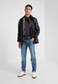Zwarte lichte jas over een donker poloshirt, lichtblauwe jeans en witte sneakers met bruine zolen. Bevat een geruite voering en een geweven riem.