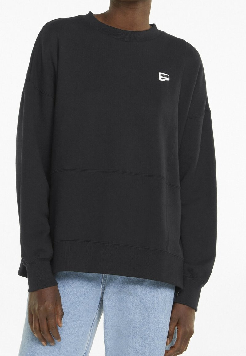 Personne portant un sweat-shirt noir Puma avec un petit logo sur la poitrine et un jean bleu clair, debout devant un fond uni.