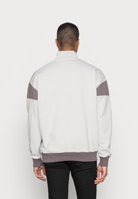 Sudadera blanca con un cuello alto, que presenta acentos en bloque de color gris y malva en los hombros y puños. Textura suave, corte relajado.