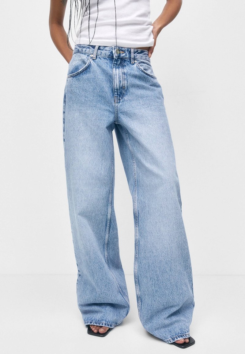 Personne portant un jean large bleu clair et un haut blanc sans manches, debout avec une main sur la hanche devant un fond uni.