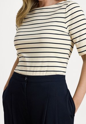 Frau trägt ein cremefarbenes kurzärmeliges Shirt mit schwarzen horizontalen Streifen und hoch taillierte dunkelblaue Hose, die Hände in den Taschen.