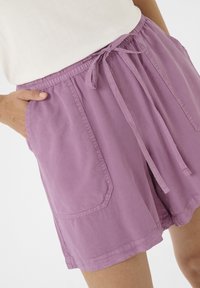 Šviesiai violetiniai šortai, pagaminti iš minkštos medžiagos, su elastine juosta, raišteliu ir šoninėmis kišenėmis.