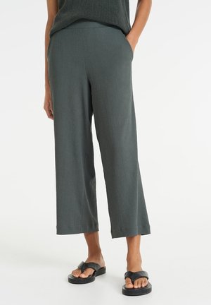 Persoon draagt losse donkergrijze cropped broek en zwarte sandaaltjes met open tenen, staande met één hand in de zak tegen een witte achtergrond.