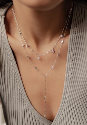 Silberne mehrschichtige Halskette mit sternförmigen Akzenten, bestehend aus einer zarten Kette in verschiedenen Längen, präsentiert vor einem strukturierten grauen Oberteil.