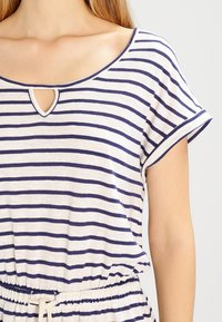 Haut rayé crème avec des rayures marine, présentant un décolleté rond avec un petit détail en forme de trou de clé et des manches courtes roulées. Taille élastique.